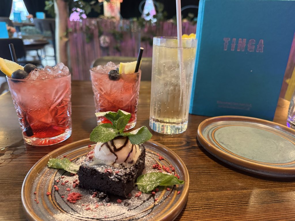 A table in a restaurant. Front centre is a plate with a slice of chocolate brownie, topped with ice cream, splashed in chocolate sauce and mint leaves. Behind the plate of chocolate brownie is a large lime and soda, and two glasses of berry vodka cocktail, with blueberries, a slice of lemon & ice cubes visible.