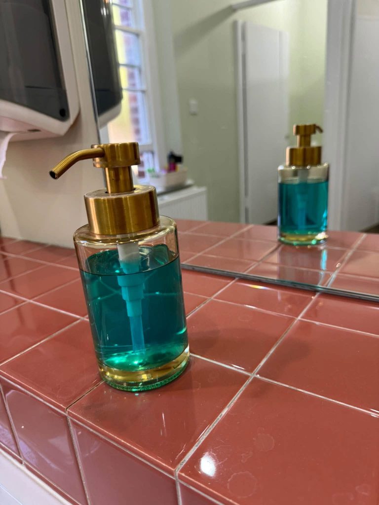 A red tiled splashback in front of a mirror. On the splashback shelf is a see through glass container, filled with bluish colour liquid soap, and a gold colour dispenser